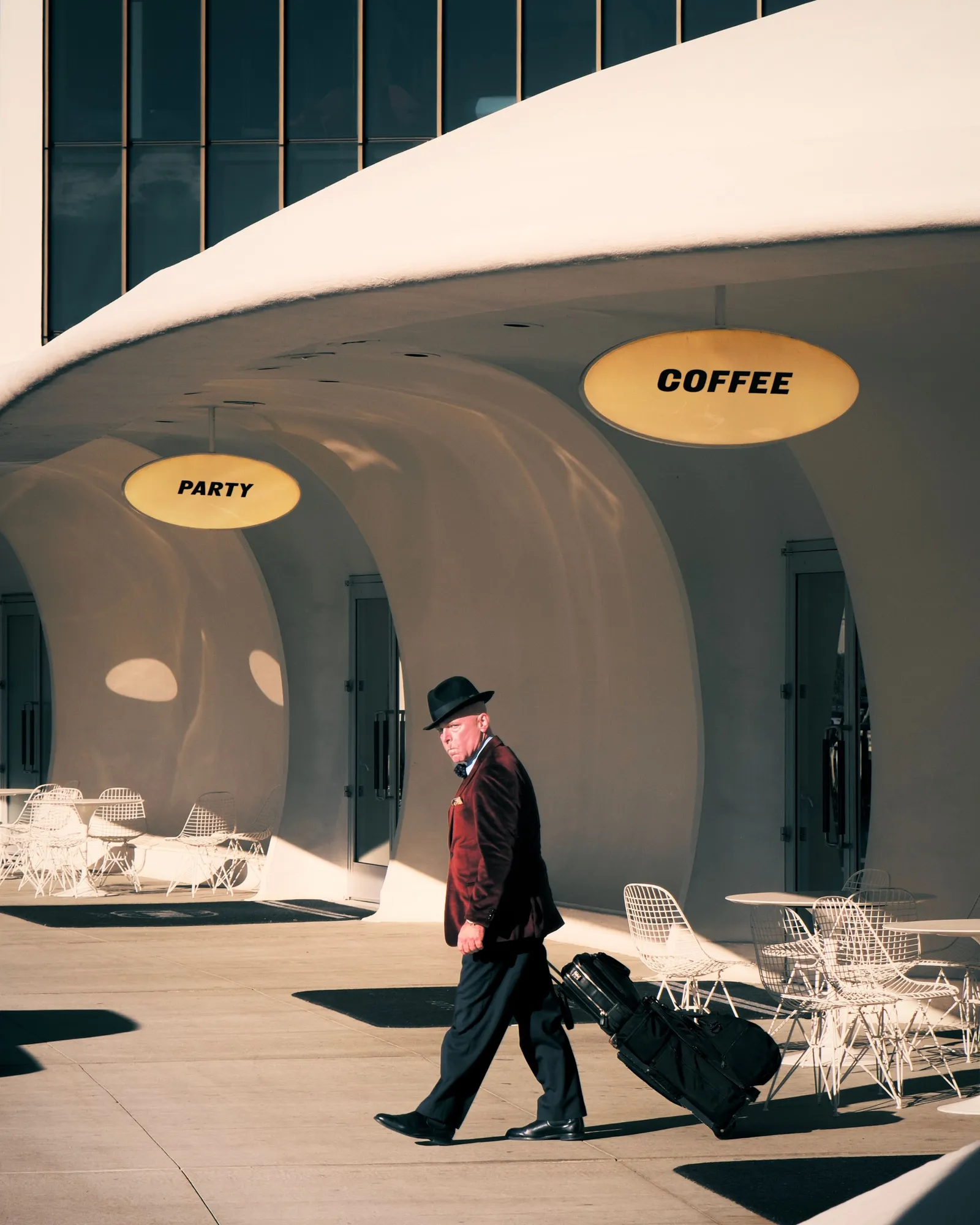 A man in a hat and velvet jacket pulling a suitcase past curved white arches, by Alex Zarnoski