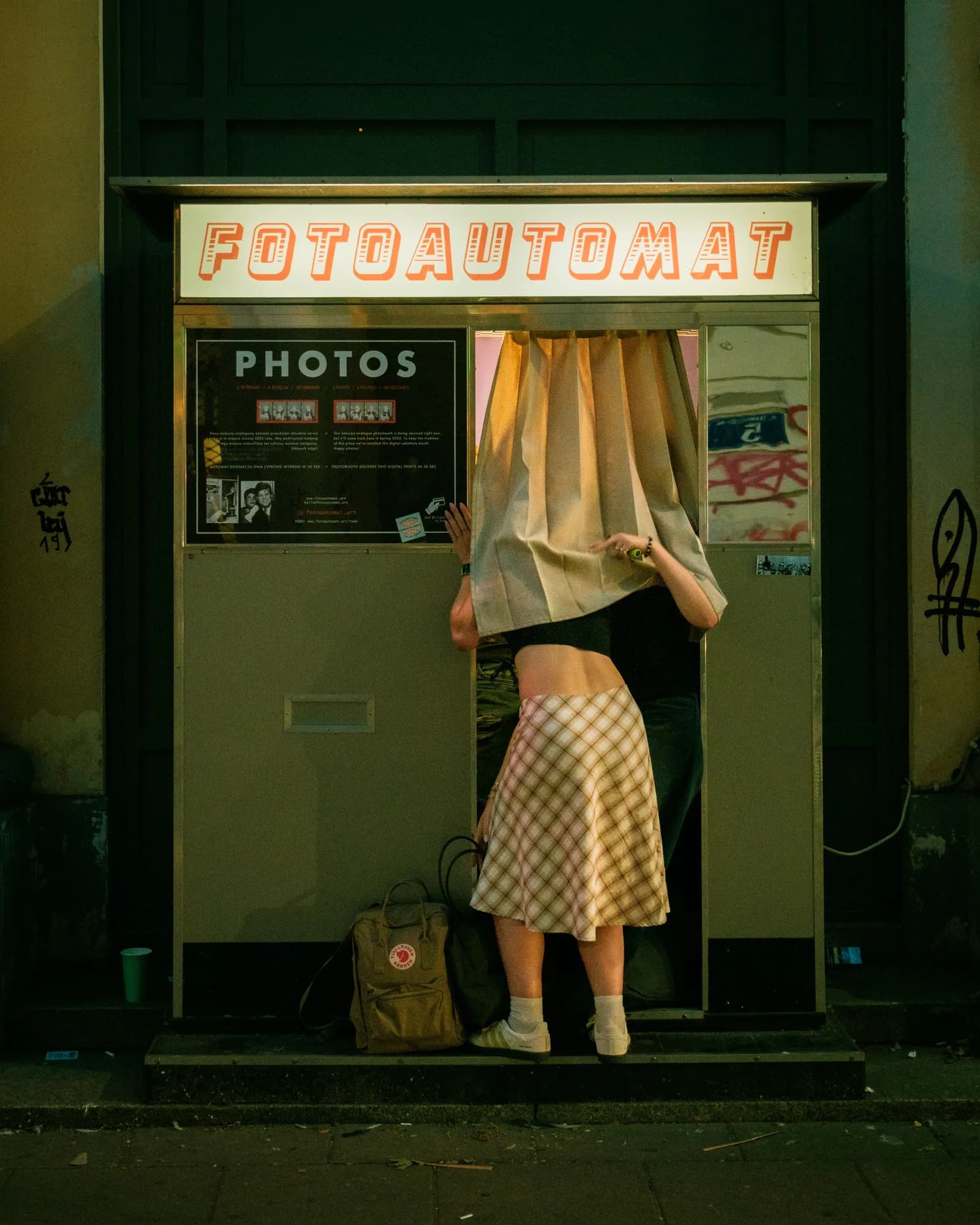 A woman leaning into a lit Fotoautomat photo booth at night, by Jon Bilous